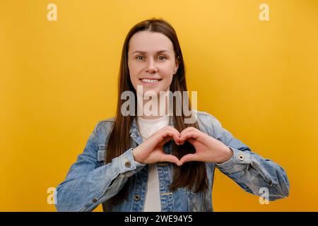 Ritratto di una giovane donna simpatica in piedi con gesto d'amore, sorridente guardando la fotocamera, mostrando il gesto a forma di cuore, indossando una giacca in denim, posando isolata Foto Stock