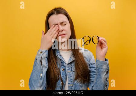 Ritratto di una giovane donna esausta e infelice degli anni '25 si toglie gli occhiali, sente dolore agli occhi, essere stanco per le lunghe ore di lavoro, indossare una giacca in denim, posare è Foto Stock