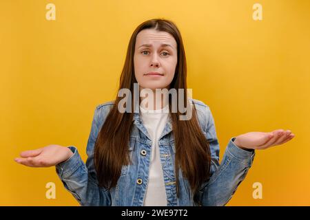 Ritratto di una giovane donna perplessa con le braccia fuori, che si stringe le spalle, dicendo, quindi, non so, indossa una giacca in denim e posa isolata su plai Foto Stock