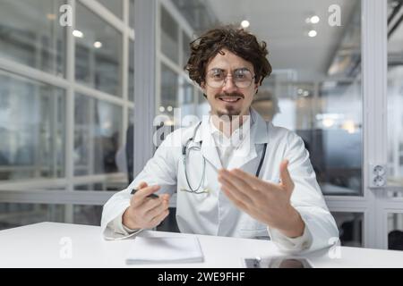 Un medico che indossa un camice bianco da laboratorio e uno stetoscopio è seduto a un tavolo all'interno di uno studio medico. Fotocamera dall'aspetto sorridente, videochiamata, utilizzo di un computer portatile, webcam Foto Stock