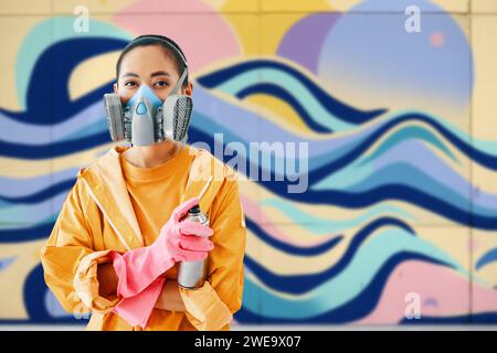 Graffiti artista di strada donna con maschera respiratoria in piedi vicino al muro con i suoi dipinti che guardano alla fotocamera con spazio per la copia. Concetto di arte urbana Foto Stock