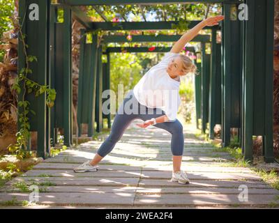 Giovane donna che conduce uno stile di vita sano e pratica yoga, esegue l'esercizio virabhadrasana, posa guerriera, si allena da sola la mattina d'estate nel parco. Concetto di benessere Foto Stock