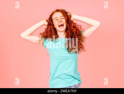 Urlante giovane donna dai capelli rossi con capelli ondulati ricci con orecchie chiuse in maglietta verde su sfondo rosa ho chiuso con tutti Foto Stock