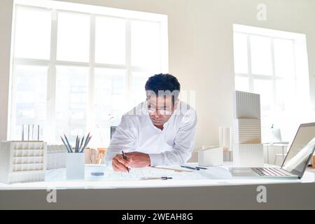 Ritratto di un architetto indiano maschio che disegna i piani presso la postazione di lavoro all'interno di un ufficio bianco, spazio per la copia Foto Stock