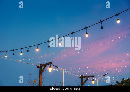 Lampadina retrò che si illumina con un cordoncino appeso decorativo al cortile per festeggiamenti al tramonto Foto Stock