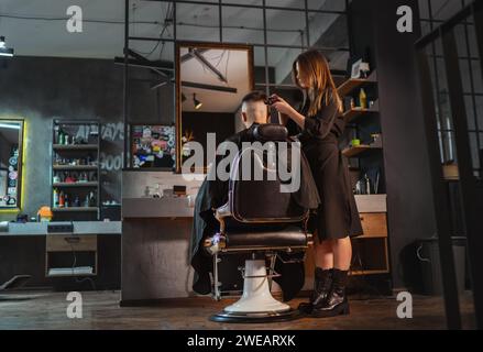 Bella giovane barbiera che crea l'acconciatura da uomo con il Clipper per capelli in stile loft moderno all'interno del barbiere. Servizi di parrucchiere locali Foto Stock