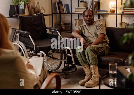 Veterano nero con disabilità seduto sul divano in posa rilassata e parlando con il terapista prendendo appunti Foto Stock