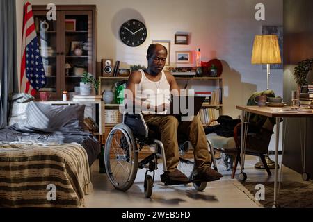 Uomo di colore con disabilità fisica seduto nella sua camera da letto e che naviga sul notebook che lavora in remoto Foto Stock