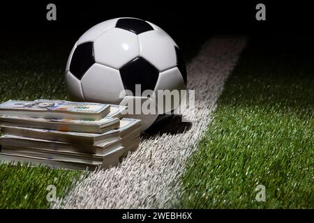 Una palla da calcio su un campo in erba con una striscia da una pila di banconote da cento dollari Foto Stock