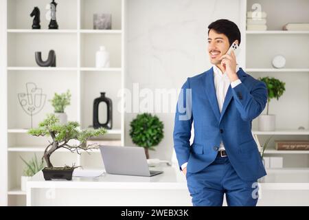 L'uomo d'affari ispanico gode di un momento spensierato durante una telefonata, la sua espressione di facilità e positività in uno spazio ufficio ben organizzato Foto Stock