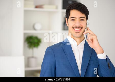 Il carismatico uomo d'affari ispanico in abito blu è impegnato in una conversazione su smartphone, messo a confronto con un ufficio moderno, combinando la tradizionale condotta aziendale e il design contemporaneo dello spazio di lavoro Foto Stock