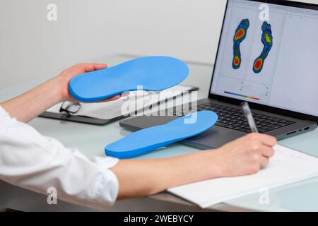 Medico ortopedista che tiene solette personalizzate in una clinica di fronte al computer portatile con un'immagine dei piedi di prova. Attività ricreative per i piedi e mezzi ortotici Foto Stock