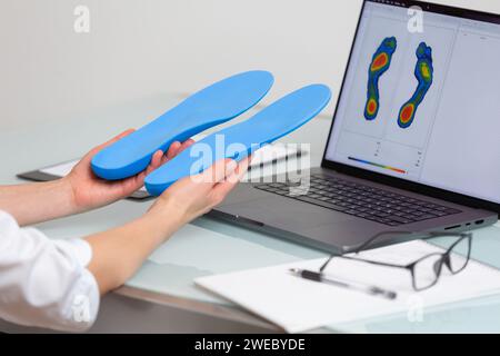 Medico ortopedista che tiene solette personalizzate in una clinica di fronte al computer portatile con un'immagine dei piedi di prova. Attività ricreative per i piedi e mezzi ortotici Foto Stock