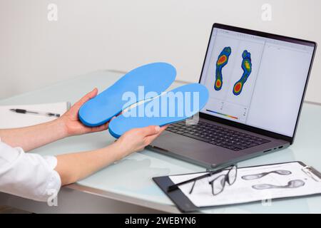 Medico ortopedista che tiene solette personalizzate in una clinica di fronte al computer portatile con un'immagine dei piedi di prova. Attività ricreative per i piedi e mezzi ortotici Foto Stock