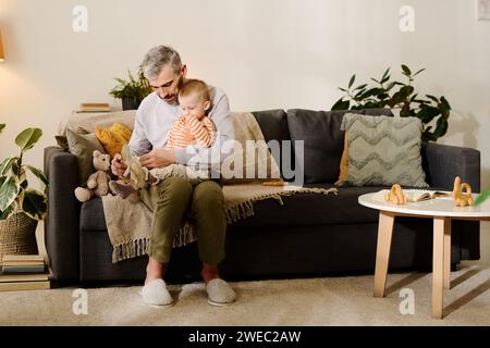 Uomo maturo in abbigliamento casual seduto sul divano in salotto e tenendo in ginocchio il bambino carino mentre si mette le scarpe da cross sui piedi Foto Stock