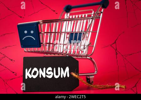 24 gennaio 2024: Carrello con smiley negativo triste, sfondo sgretolante ed etichetta con l'iscrizione: Konsum FOTOMONTAGE *** Einkaufswagen mit einem traurigen negativen Smiley, bröckelnden Hintergrund und und einem Etikett mit Aufschrift: Konsum FOTOMONTAGE Foto Stock