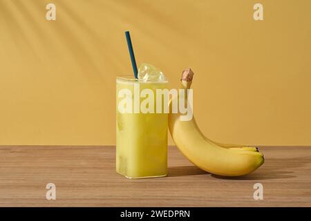 Vista frontale di un bicchiere di succo di banana e di una banana gialla matura su un tavolo di legno su sfondo giallo con ombra di foglie di palma. Scena per pr. Pubblicitario Foto Stock