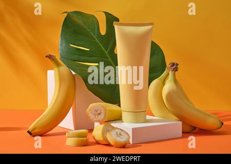 Un tubo cosmetico giallo su podio bianco, decorato con banane fresche e foglie di monstera della giungla su sfondo giallo con ombra di foglie. Scena di mockup Foto Stock