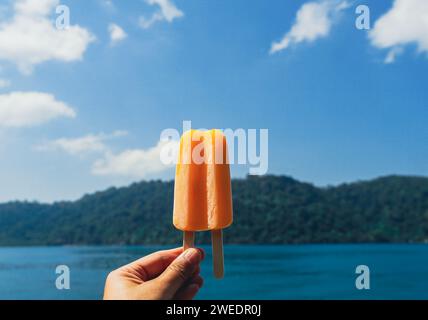 Gelati arancioni su sfondo blu. Ghiacciolo arancione nella mano di una donna. Mano femminile che tiene il ghiacciolo arancione che scioglie su sfondo blu cielo in somme Foto Stock