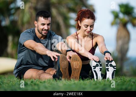 Felice giovane coppia adulta che si allunga per terra dopo aver corso insieme nel parco. Sorridendo soddisfatto atletico maschile e femminile seduto nell'erba Foto Stock