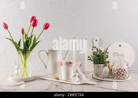 Natura morta elegante per le vacanze di Pasqua. Caffettiera bianca su vassoio bianco in ceramica con tazza e personaggi coniglietto pasquale. vaso di fiori e candele Foto Stock