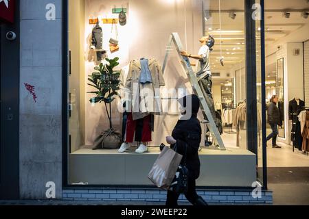Una donna si trova su una scala a pioli all'interno di un negozio di abbigliamento mentre riorganizza la vetrina in Oxford Street, Londra, Inghilterra, Regno Unito Foto Stock