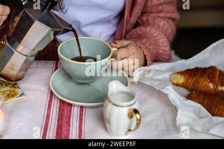 Primo piano di mani che versano un espresso caldo da una moka in una tazza, con un delizioso croissant in attesa. Foto Stock