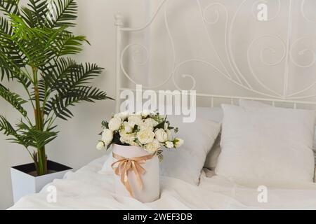 Mattina di compleanno con un regalo di rose bianche fresche a letto. Elegante camera da letto bianca con bouquet fresco e piante tropicali Foto Stock