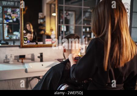 Giovane barbiera che crea un taglio di capelli tagliati da uomo utilizzando il tagliacconciatura di fronte allo specchio barbiere in stile moderno. Servizio di cura dei capelli sma locale Foto Stock