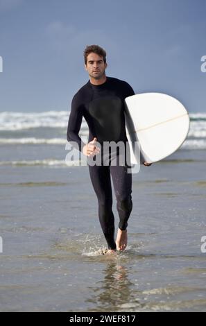 Uomo, ritratto e spiaggia sull'acqua con tavola da surf per onde, esercizio fisico o hobby all'aperto in fitness, surf o pratica. Persona maschile o surfista con tavola Foto Stock