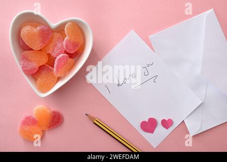 Nota scritta i Love You su carta bianca su sfondo rosa con matita e contenitore in ceramica bianca con dolci a forma di cuore. Vista dall'alto. Foto Stock