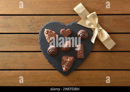 Assortimento di cioccolatini su piastra in ardesia a forma di cuore con fiocco dorato al centro su tavola in legno a doghe. Vista dall'alto. Foto Stock