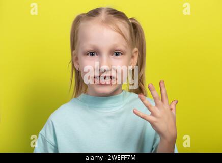 Una bambina di sette anni con una bocca sporca di cioccolato su sfondo giallo. Concetto di bambini con golosi, livelli di zucchero nel sangue, diabete e. Foto Stock
