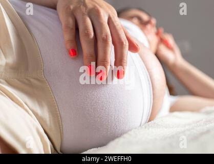 una ragazza incinta giace sul letto e le tiene lo stomaco. Concetto di dolore e disagio addominale. Esacerbazione di malattie croniche nelle donne incinte. Foto Stock