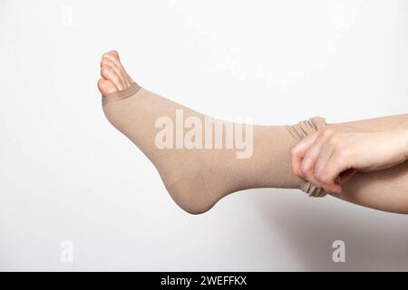 Una donna mette su calze di compressione per gambe con vene varicose. Dolore e gonfiore alle gambe, flebologia. Sfondo bianco, primo piano. Foto Stock