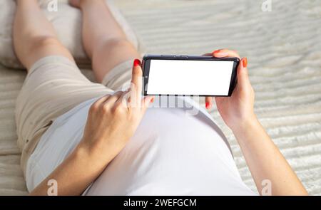 Una ragazza incinta è sdraiata sul letto e tiene uno smartphone tra le mani. Concetto di applicazioni negli smartphone per donne in gravidanza, numero di contrazioni Foto Stock