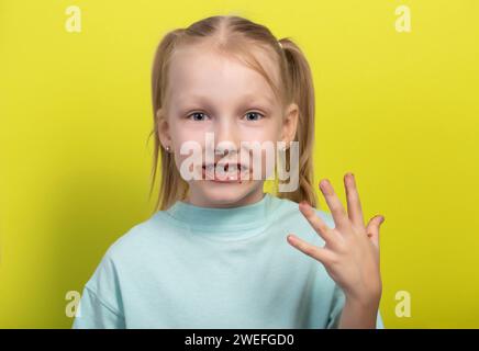 Una bambina di sette anni con una bocca sporca di cioccolato su sfondo giallo. Concetto di bambini con golosi, livelli di zucchero nel sangue, diabete e. Foto Stock