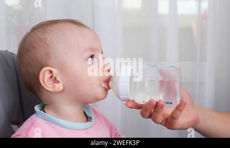 La mano di una madre dona acqua a un bambino da bere da un biberon mentre allatta il porridge. Un bambino di un anno beve acqua. Spazio di copia per il testo Foto Stock