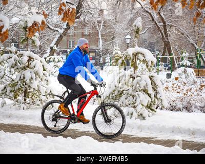 Un uomo gioioso cavalca una bicicletta in un parco invernale tra alberi innevati. Ciclista barbuto in una giacca blu.. Stile di vita attivo Foto Stock
