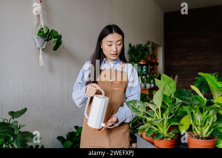 Serena ragazza coreana che nutre fiori in vaso in un laboratorio o in una casa, innaffiando delicatamente ogni fiore con una delicata lattina. Foto Stock