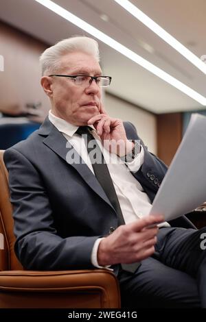 Uomo d'affari anziano in abbigliamento da formalwear seduto in poltrona e attento a leggere informazioni su carta Foto Stock
