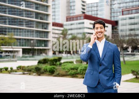 Un uomo d'affari ispanico è impegnato in una conversazione sul suo cellulare, sorridendo sottilmente con un'aria di successo mentre si trova di fronte a un paesaggio urbano, incarnando il moderno professionista del multitasking. Foto Stock