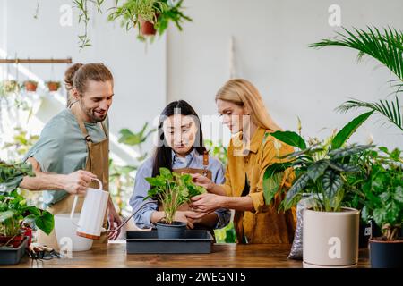 Giornata di lavoro impegnativa al fioraio. Colleghi di alberi, donna asiatica, giovane uomo e padrona bionda di successo di mezza età insieme. Impianto di irrigazione maschile. Foto Stock