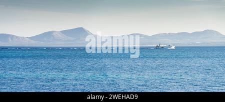 Un peschereccio a strascico attraversa le acque blu cobalto, creando un sentiero spumeggiante, sullo sfondo sereno di lontane montagne sotto un Foto Stock