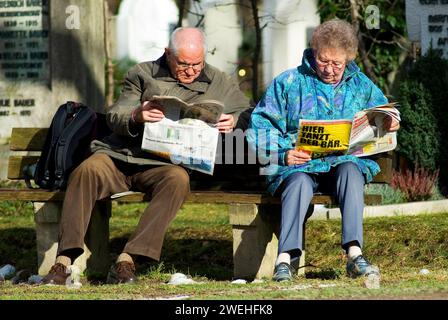 Una coppia anziana è seduta su una panchina sotto il sole primaverile leggendo un giornale, si possono vedere le parole "qui l'orso balla” Foto Stock