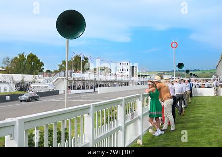 Spettatori al circuito automobilistico Goodwood Revival. Giovane donna con abito verde a pieghe, occhiali da sole bianchi e fascia a testimonianza della moda retrò. Foto Stock
