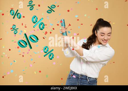 Offerta scontata. Felice giovane donna che fa saltare in aria Party popper su sfondo beige scuro. Coriandoli e segni di percentuale nell'aria Foto Stock