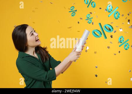 Offerta scontata. Felice giovane donna che fa saltare in aria Party popper su sfondo dorato. Coriandoli e segni di percentuale nell'aria Foto Stock