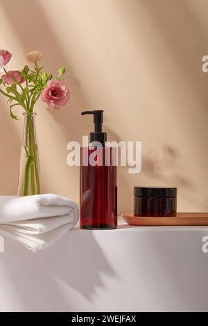 Set di biberon per la cura del corpo color ambra senza etichetta, asciugamano di cotone e un vaso di fiori rosa su sfondo beige. Ombra naturale dell'albero sul muro. Scena mockup f Foto Stock