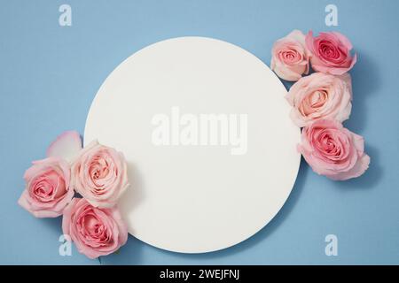 Design minimalista, vista dall'alto delle rose delicate disposte su sfondo blu con podio bianco rotondo vuoto per messaggi promozionali o branding Foto Stock